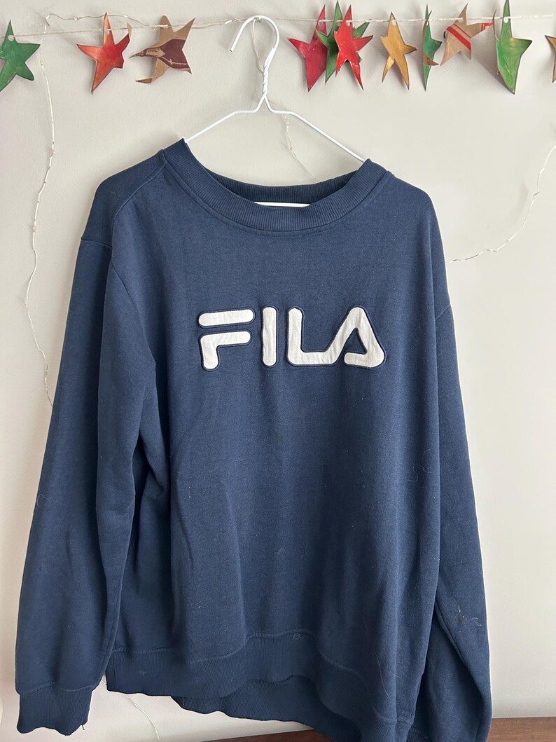 May include: A navy blue long-sleeve sweatshirt featuring a crew neckline and ribbed cuffs. The front of the garment displays the word FILA in large, raised, white block letters with a dark blue outline, centred on the chest area.