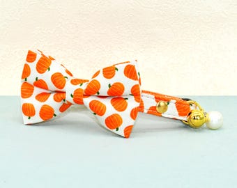 May include: A white cat collar with an orange pumpkin pattern and a matching bow tie. The collar has a gold-colored bell and a white pearl charm. The bow tie and collar are made of fabric.