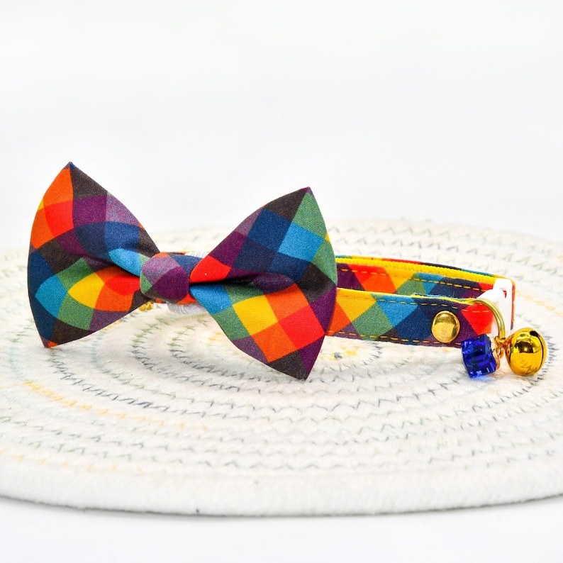 May include: A colorful plaid bow tie cat collar with a bell and a blue charm. The collar is yellow, red, orange, blue, green, and purple.