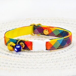 May include: A colorful cat collar with a yellow backing, a white buckle, and a blue bell charm. The collar has a plaid pattern with red, orange, yellow, green, blue, and purple squares.