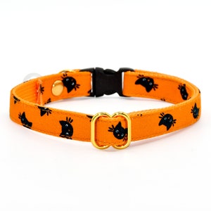 May include: Orange cat collar with black cat faces printed on it. The collar has a gold metal buckle and a black plastic buckle.