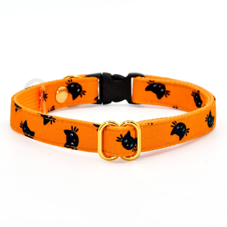 May include: Orange cat collar with black cat faces printed on it. The collar has a gold metal buckle and a black plastic buckle.