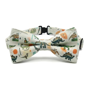 Peut inclure: Nœud papillon crème avec un motif répété de dinosaures verts, oranges et bleus. Le nœud papillon a un collier et une boucle noirs réglables. Le motif comprend diverses espèces de dinosaures.