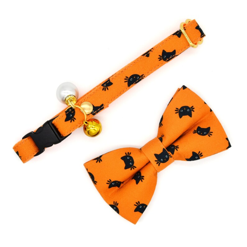 May include: Orange cat-themed pet collar with a bell and a matching bow tie. The collar has a black buckle and the bow tie has a black cat print.