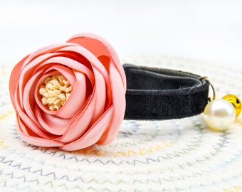 May include: A pink fabric flower with a black velvet collar and a pearl and gold bell charm.