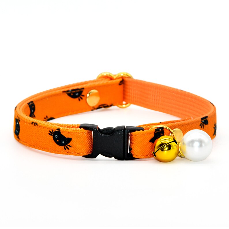May include: Orange cat collar with a black cat face pattern, a gold bell, and a white pearl charm.