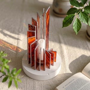 May include: A candle holder featuring vertical stained glass panels in shades of orange and brown, arranged in a spiral pattern on a circular white concrete base. A single white pillar candle sits in the center of the glass structure.