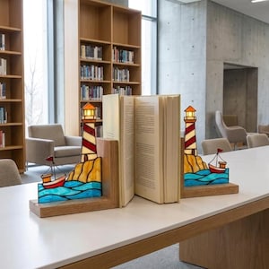 Könnte beinhalten: Ein Paar L-förmige Buchstützen aus Holz mit Buntglas-Leuchtturmmotiven in Rot und Weiß, gelben Felsensockeln und kleinen rot-weißen Segelbooten auf blauen Wellen, die ein aufgeschlagenes Hardcover-Buch auf einem weißen Tisch stützen.