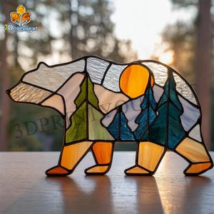 May include: Stained glass bear figurine with a mountain and forest scene. The bear is made of various colors of glass, including orange, green, blue, and white. The sun is a bright yellow circle. The text "3DPrintyourart" is visible.
