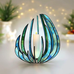 May include: A stained glass candle holder featuring curved, flame-like panels in shades of blue and green with black metal framing. A single white pillar candle sits in the centre, emitting a soft glow from its lit wick within the structure.