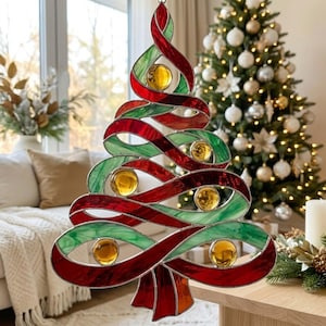 May include: A stained glass Christmas tree ornament featuring swirling red and green ribbons with embedded amber-colored glass spheres. The design is constructed with dark metal solder lines forming a continuous, flowing, abstract tree shape.