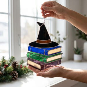 Puede incluir: Un atrapasoles de vidrieras que muestra una pila de tres libros en azul, verde y morado con un sombrero de bruja negro encima. La pieza está construida con líneas de soldadura de metal oscuro e incluye una cadena plateada para colgarla.