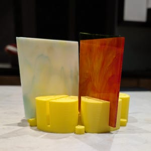 May include: Abstract art piece featuring a combination of materials and colors. The composition includes a translucent, marbled white panel, a vibrant orange and red glass panel, and several yellow cylindrical supports.
