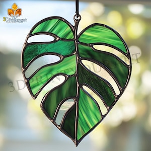May include: A stained glass window hanging features a green monstera leaf with a black metal frame. The leaf is made of multiple pieces of glass, creating a textured look.