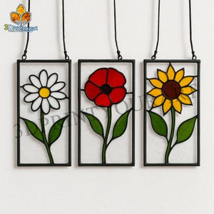 May include: Three stained glass panels with floral designs. A daisy with white petals and a yellow center, a red poppy, and a yellow sunflower with a brown center. Each panel is framed in black and has a hanging cord.