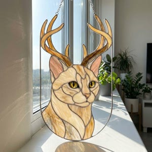 Puede incluir: Un atrapasoles de vidrieras que muestra una cabeza de gato con grandes astas de ciervo. La pieza está hecha de segmentos de vidrio crema, bronceado y marrón con ojos amarillos, encerrada en un marco de metal fino y colgada de una cadena plateada.