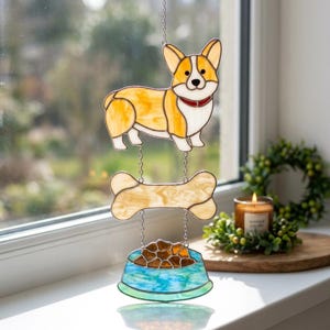 Puede incluir: Un atrapasoles de vidrieras que muestra un perro Corgi de color canela y blanco, un hueso beige y un cuenco azul lleno de comida marrón. Las tres piezas están unidas por cadenas de metal plateado, creando un adorno vertical para colgar.