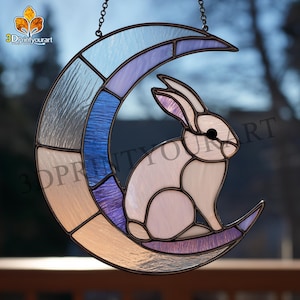 Peut inclure: Suncatcher en vitrail représentant un lapin blanc assis sur un croissant de lune. La lune est segmentée avec des nuances de bleu, de violet et de beige. Le capteur solaire est suspendu par une chaîne.