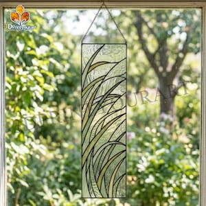 Op de afbeelding: Een rechthoekig glas-in-loodpaneel met een ontwerp van gestileerde groene en gele grassprieten. Het paneel is zwart omlijst en hangt aan een ketting. De achtergrond toont een vage buitenscène.