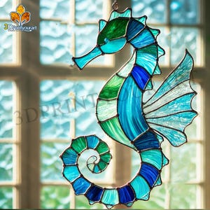 Op de afbeelding: Een gebrandschilderd zeepaardje in een blauwe, groene en turquoise kleurstelling. Het zeepaardje hangt voor een raam.