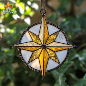 Puede incluir: Un adorno de brújula de vidrio emplomado con un diseño de estrella dorada y blanca. El adorno está colgado de una cuerda negra.