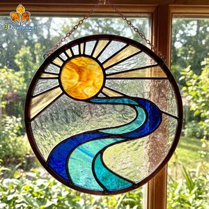 May include: A circular stained glass suncatcher featuring a yellow sun with radiating beams above a winding river rendered in shades of blue and teal. The piece is framed in dark metal and suspended by a thin metal chain for window display.