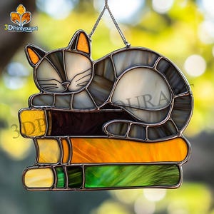 May include: A stained glass cat sleeping on a stack of books. The cat is grey and white with black stripes. The books are yellow, orange and green.