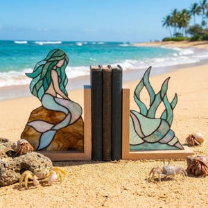 Puede incluir: Un par de sujetalibros de vidrieras que representan una sirena sentada sobre una roca y una concha marina con plantas acuáticas. Las piezas están montadas sobre bases de madera clara en forma de L, con dos libros de tapa oscura entre los paneles.