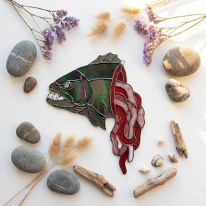 Könnte beinhalten: Ein Buntglas-Piranha-Ornament mit irisierenden dunkelgrünen Schuppen, einem scharfzahnigen Kiefer und fließenden roten sowie durchscheinenden rosa Glaselementen, die vom Körper herabhängen, verbunden durch dunkle Metalllötlinien.