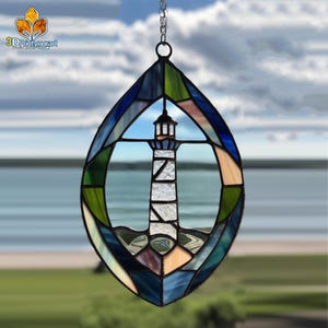 May include: A stained glass suncatcher depicting a lighthouse within a blue and green oval frame. The suncatcher is suspended from a silver chain.
