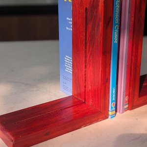 Puede incluir: Un par de sujetalibros de madera roja hechos a mano que sostienen libros infantiles. Los sujetalibros son rectangulares con una sección vertical y una base. Los libros incluyen "Robinson Crusoe" y "Jack and the Beanstalk".