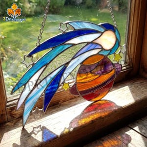 May include: Stained glass celestial art piece depicting a comet with a planet. The comet features shades of blue, green, and white, with star accents. The planet is a mix of orange, purple, and red. The piece hangs from a chain.