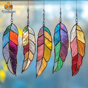 Può includere: Cinque acchiappasole a forma di piuma in vetro colorato appesi a una corda. Le piume sono fatte di diversi pezzi di vetro colorato e hanno uno schema di colori arcobaleno.