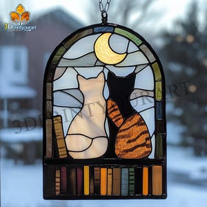 Op de afbeelding: Een hangend glas-in-loodraam met twee katten die naar een halve maan kijken. De katten zitten op een plank met boeken. Het raam is omlijst met zwart metaal.