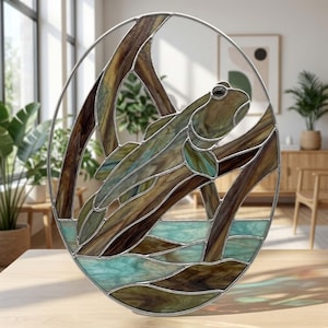 May include: An oval stained glass panel featuring a brown and green fish swimming among brown branches above light blue water. The glass pieces are held together by silver metal lines, creating a detailed aquatic scene within a circular frame.