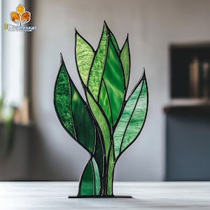 May include: A stained glass sculpture of a plant with green leaves. The leaves are made of multiple pieces of glass, creating a textured look. The sculpture is mounted on a black metal base.