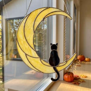 May include: A stained glass suncatcher featuring a yellow crescent moon with a black cat silhouette sitting on the lower curve. The cat is connected to the upper moon section by two thin metal chains, creating a swing-like appearance.