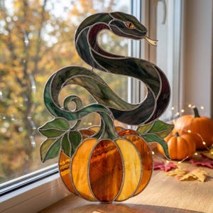 Può includere: Un acchiappasole in vetro colorato raffigurante un serpente verde scuro avvolto con occhio e lingua gialli, posizionato sopra una zucca arancione brillante con foglie in vetro verde. L'oggetto è realizzato con linee di saldatura in metallo.