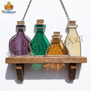 Può includere: Una decorazione da parete in vetro colorato con quattro boccette di pozione nei colori viola, verde, ambra e crema, ciascuna con tappo in sughero e bordo in metallo. Le boccette poggiano su una mensola in legno sospesa da una catena metallica.
