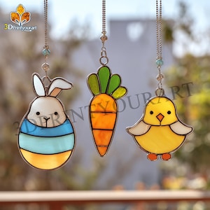 Puede incluir: Tres decoraciones de Pascua de vidrieras: un conejito, una zanahoria y un pollito. El conejito es blanco con rayas azules y amarillas. La zanahoria es naranja con hojas verdes. El pollito es amarillo con patas naranjas.