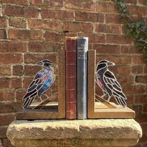 Puede incluir: Un par de sujetalibros con cuervos de vidrieras de colores con plumaje iridiscente en tonos violeta y azul, posados sobre ramas de madera. Los cuervos están montados sobre bases rectangulares de madera y sostienen dos libros antiguos.