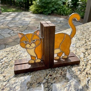 Puede incluir: Una figura de gato de vidrieras en tonos naranja y crema, dividida en dos partes por un bloque de madera marrón oscuro central. El gato tiene una expresión facial gruñona y una cola curva, montada sobre una base rectangular de madera.