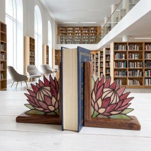 May include: A pair of wooden bookends featuring stained glass inserts shaped like protea flowers with deep pink and light green petals. The bookends are positioned on either side of a single blue hardcover book with gold lettering on the spine.