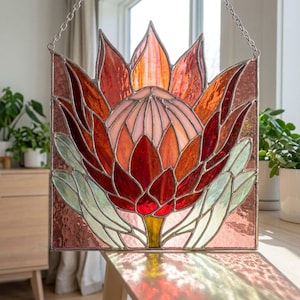 May include: A stained glass panel featuring a central protea flower in shades of deep red, orange, and soft pink. The flower is surrounded by pale green leaves and set against a textured, dusty rose background, all held together by metal cames.