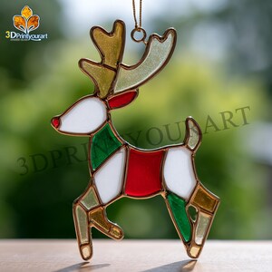 Puede incluir: Adorno navideño de reno de vidriera. El reno presenta una nariz roja, cuerpo blanco y detalles verdes y rojos. Las astas son doradas y el adorno cuelga de una cadena dorada. El logotipo "3DPrintyourart" está en la esquina superior izquierda.