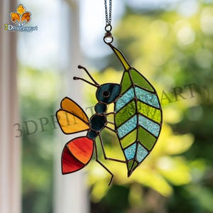 Könnte beinhalten: Buntglas-Kunstwerk, das eine Ameise mit einem Blatt darstellt. Die Ameise hat einen roten, orangefarbenen und gelben Körper mit blauen und schwarzen Akzenten. Das Blatt ist grün und blau. Aufgehängt an einer Kette.