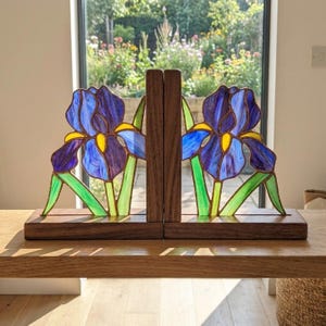 May include: A pair of bookends featuring stained glass iris flowers in shades of deep blue, purple, and yellow with green stems. Each glass panel is set into a rectangular dark brown wooden base, creating a decorative support for books.