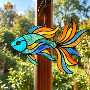 May include: A stained glass fish suncatcher with a vibrant design. The fish features a turquoise head, a blue body, and orange and yellow fins. The suncatcher is hanging in front of a window, with the text "3DPrintyourart" in the upper left corner.