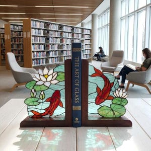 May include: A pair of dark wood bookends featuring stained glass panels with red koi fish, white lotus flowers, and green lily pads. The panels flank a blue book titled The Art of Glass, creating a decorative display on a white wooden surface.