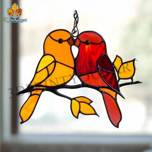 Pode incluir: Um vitral pendurado apresenta dois pássaros empoleirados em um galho com folhas. Os pássaros estão se beijando e são vermelhos e amarelos.
